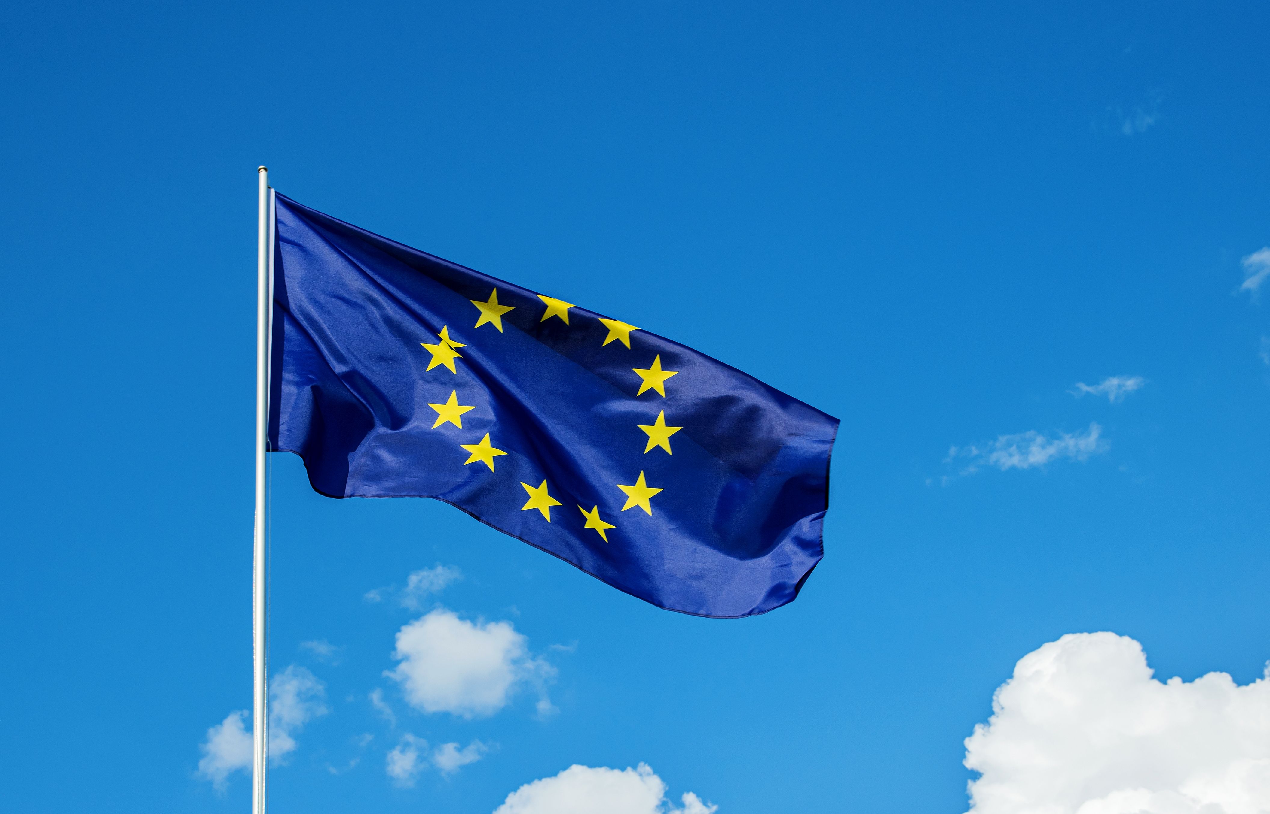 Bandeira da União Europeia tremulando ao vento em um mastro branco, com um fundo de céu azul e algumas nuvens.