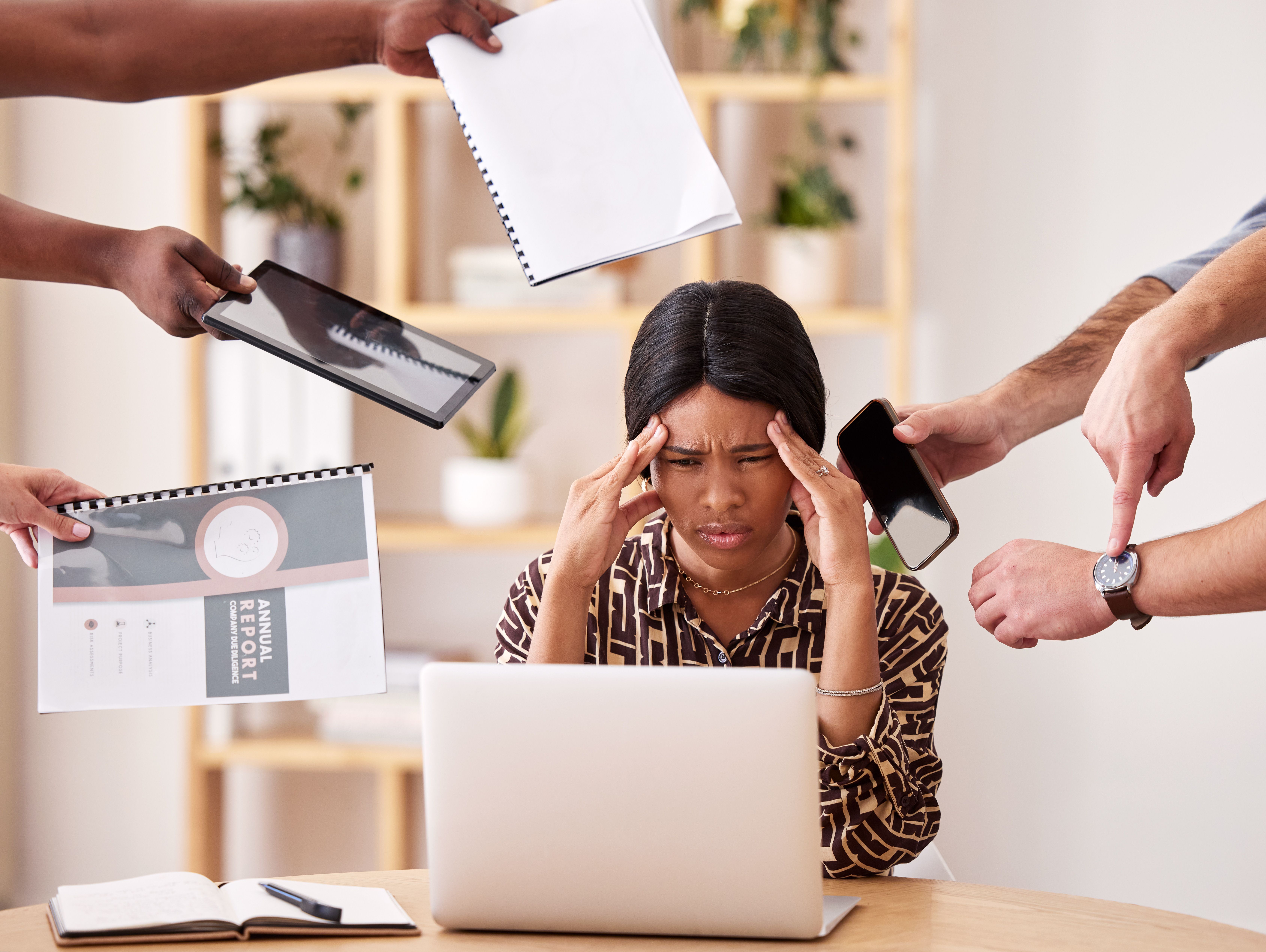Mulher estressada em frente a um notebook, pressionada por várias mãos ao redor que entregam documentos, apontam para o relógio e mostram dispositivos eletrônicos, simbolizando a sobrecarga de tarefas. 