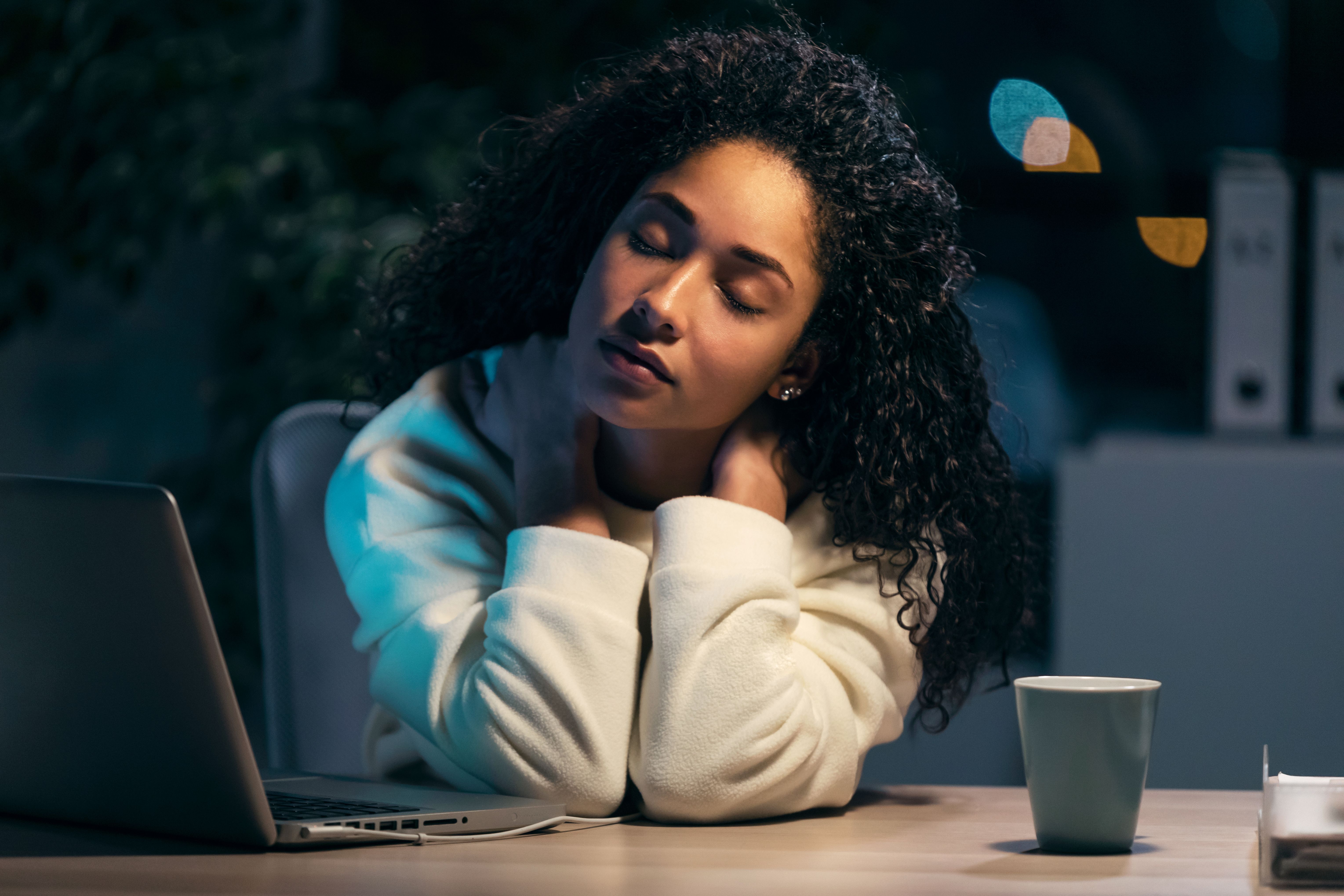 Mulher visivelmente cansada, com os olhos fechados e mãos no pescoço, sentada em frente a um laptop durante a noite. 