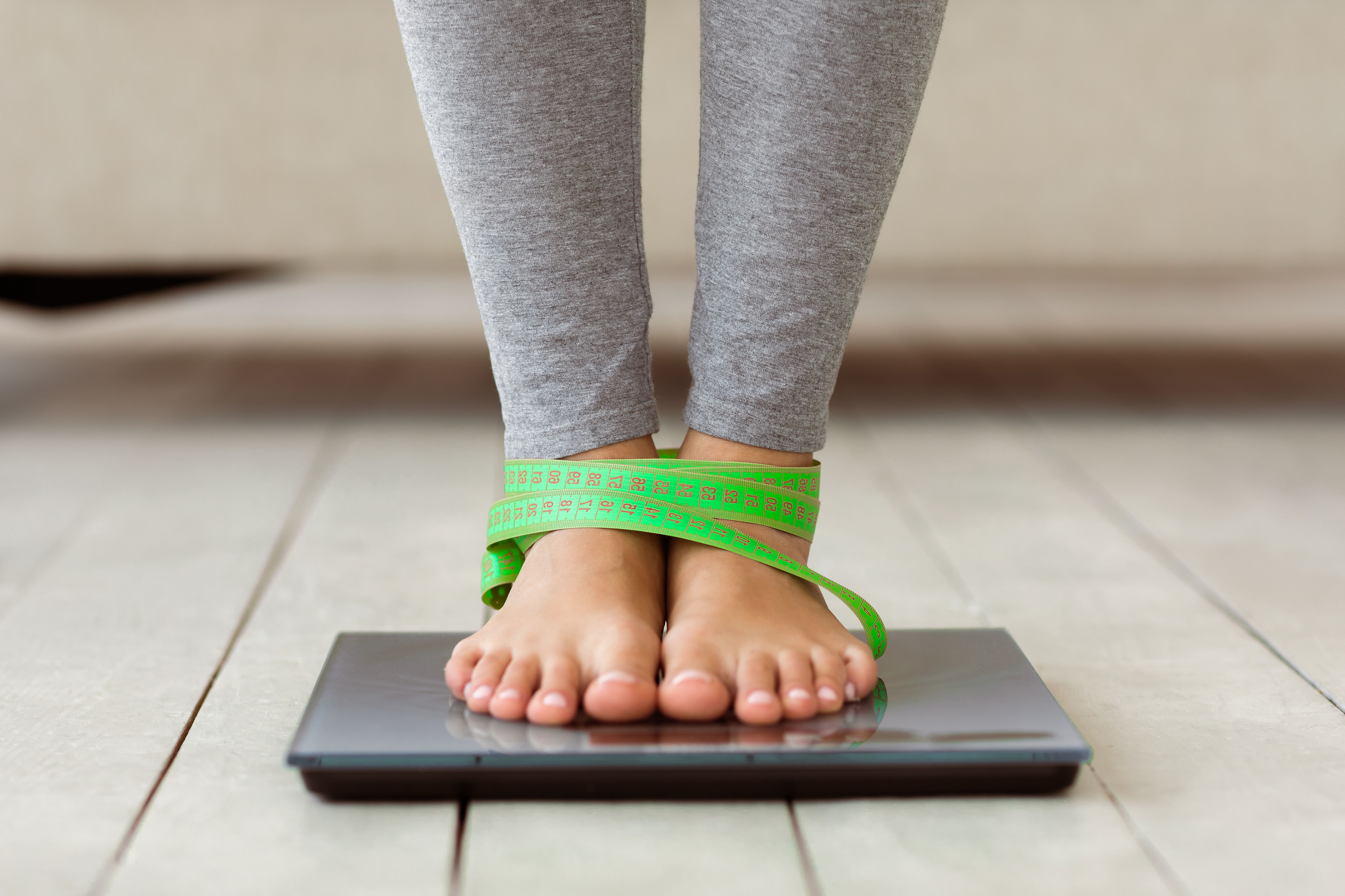 Pessoa de leggings cinza em pé sobre uma balança digital com uma fita métrica verde amarrada nos pés.