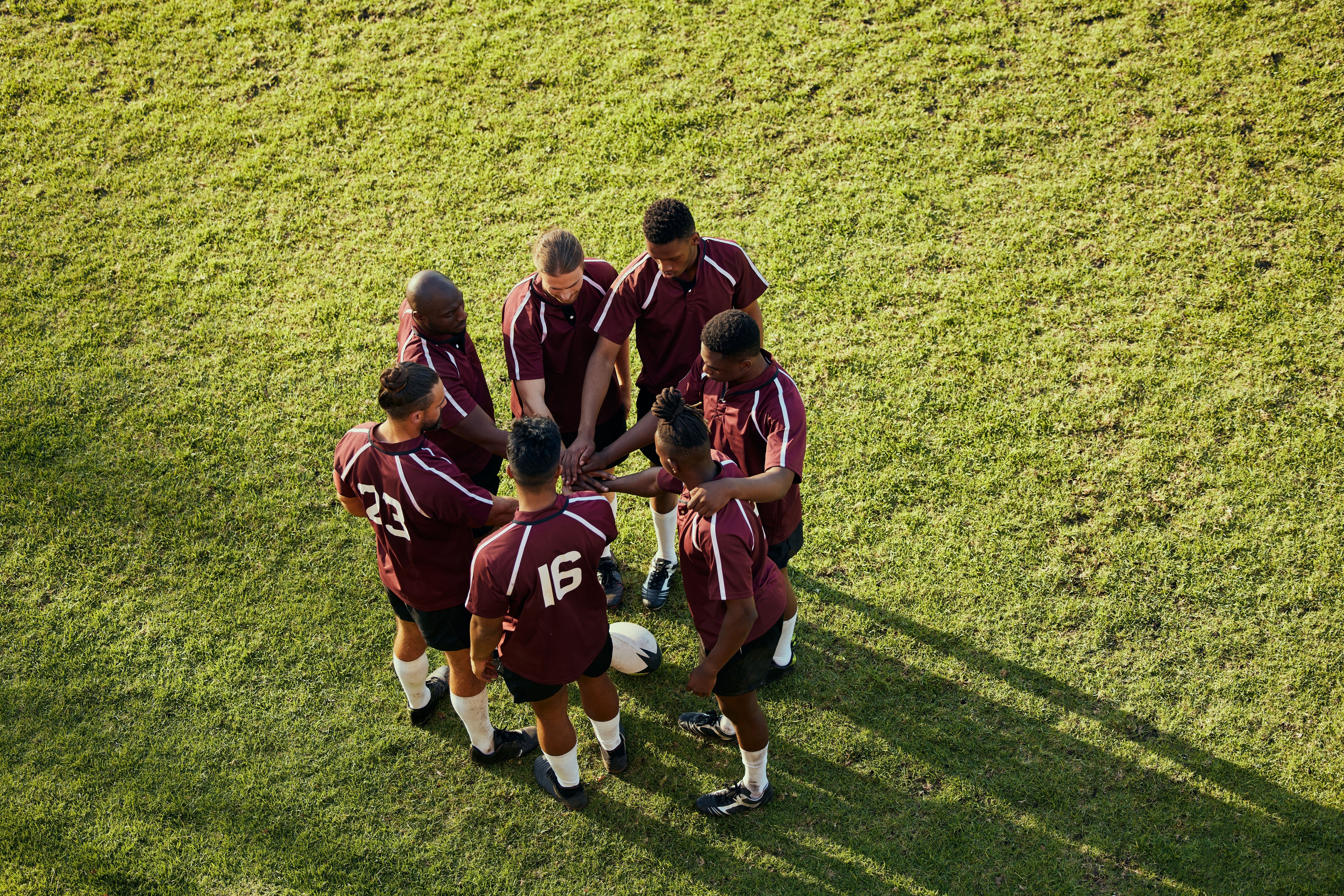 Seis jogadores de futebol, vestindo uniformes bordô com detalhes brancos, estão reunidos em círculo no campo gramado. 