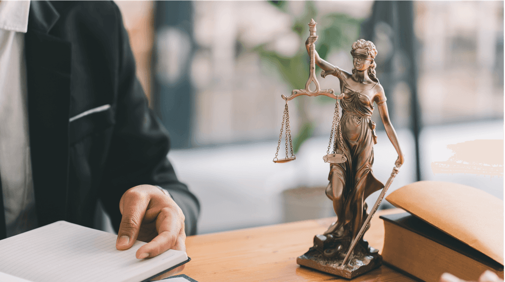 Estátua da Justiça ao lado de um advogado com livro e documentos sobre a mesa.