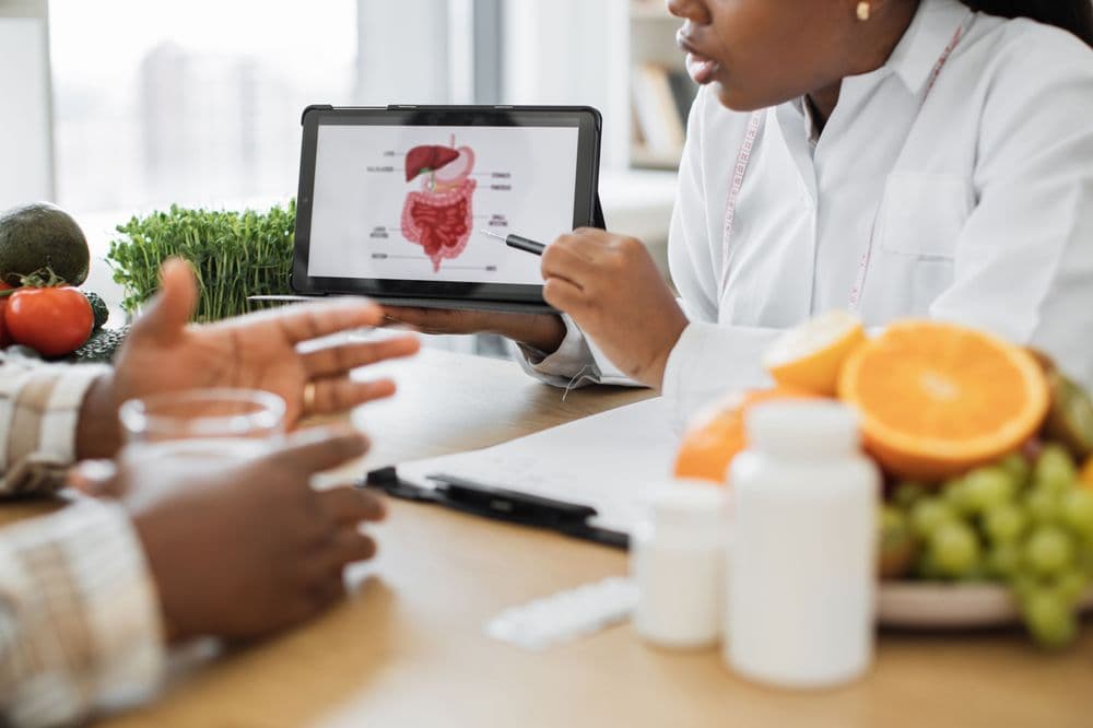 Uma médica ou nutricionista segura um tablet com uma ilustração do sistema digestivo humano, enquanto orienta um paciente em consulta.