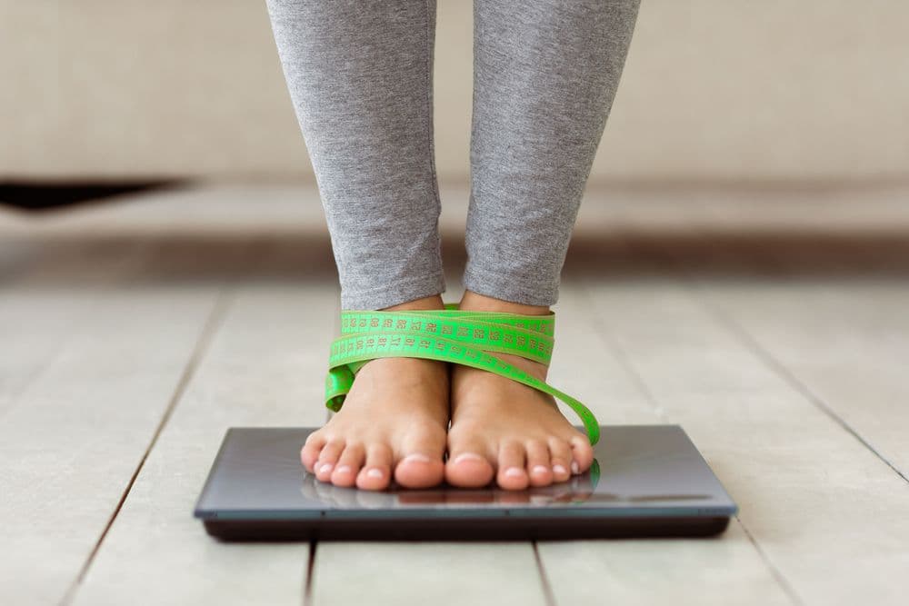 Pessoa de leggings cinza em pé sobre uma balança digital com uma fita métrica verde amarrada nos pés.