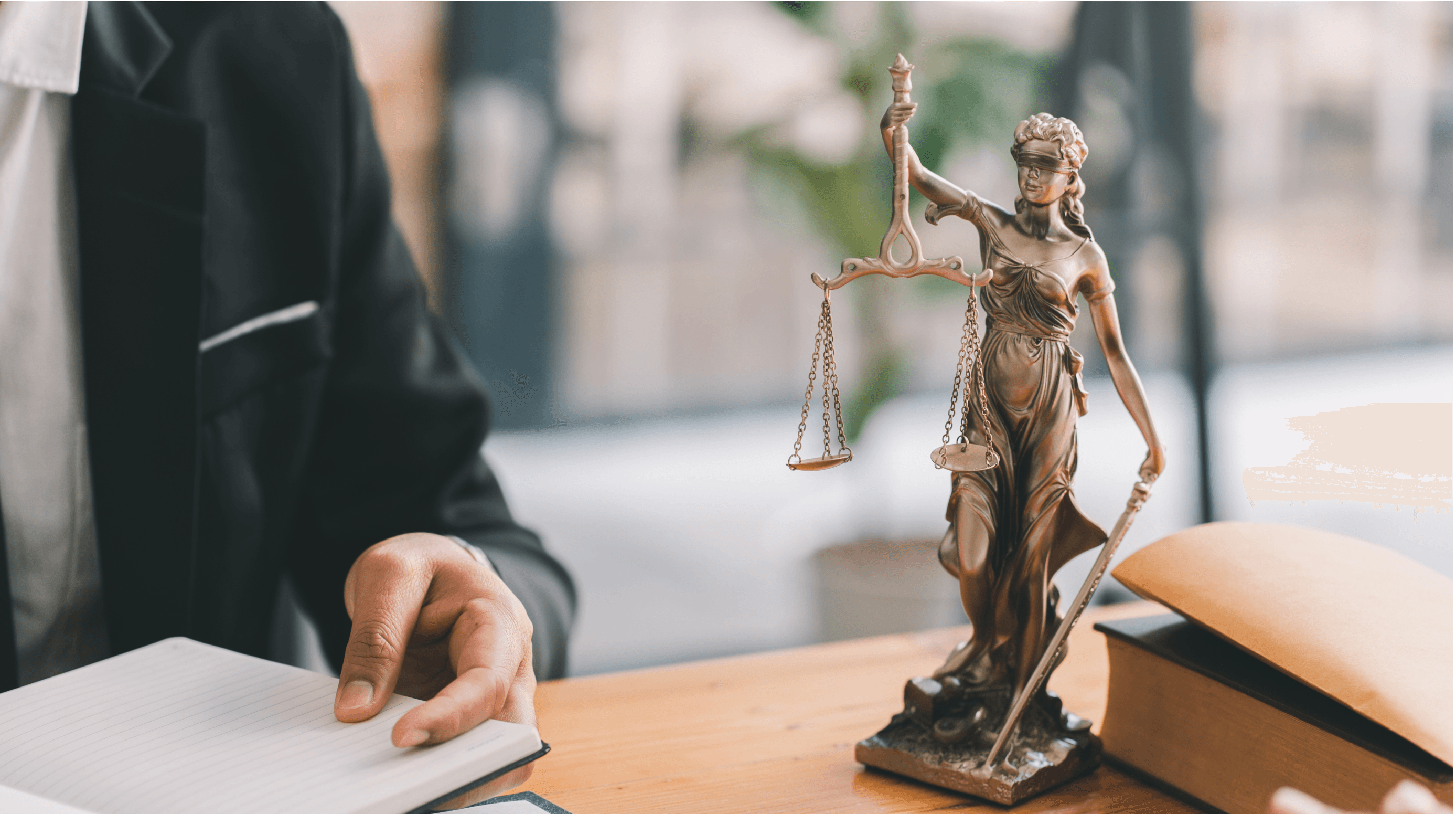 Uma estátua da Justiça com balança e espada está posicionada sobre uma mesa ao lado de um advogado, que segura um caderno aberto. Ao fundo, há um envelope e um livro jurídico.