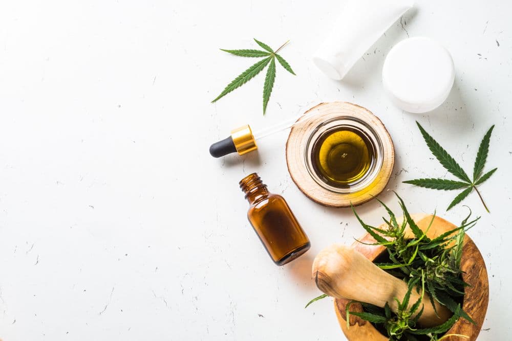 Óleo de cannabis medicinal em frasco com conta-gotas, folhas de cannabis e recipiente cosmético sobre fundo claro.