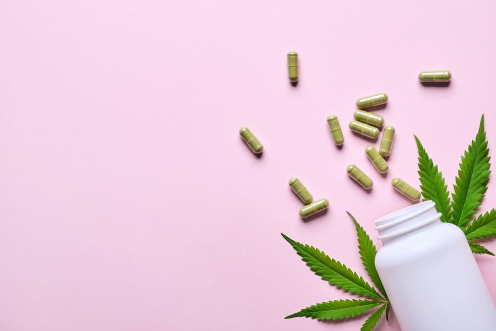 Cápsulas de CBD ao lado de folhas de cannabis em um fundo rosa.