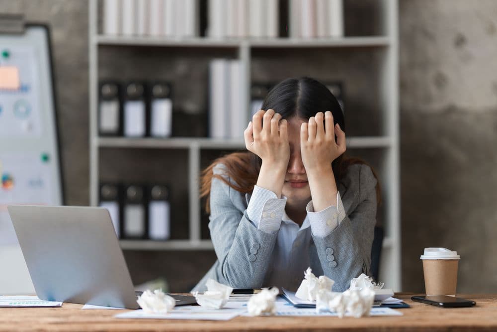 Mulher com sinais de estresse e burnout sentada à mesa de trabalho, com a cabeça apoiada nas mãos em um escritório.