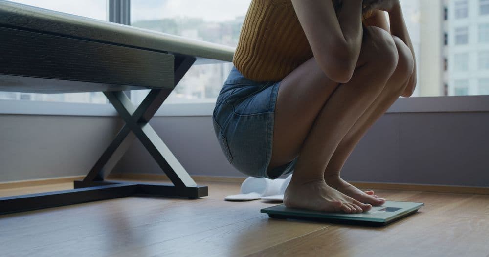 Mulher em pé sobre uma balança em ambiente doméstico, representando a relação com o peso corporal e transtornos alimentares.