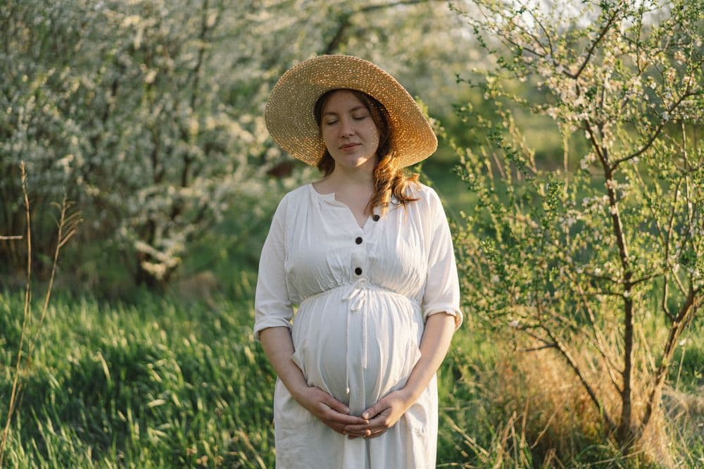 Mulher grávida em um campo verde, usando chapéu de palha e vestido branco, representando maternidade e bem-estar.
