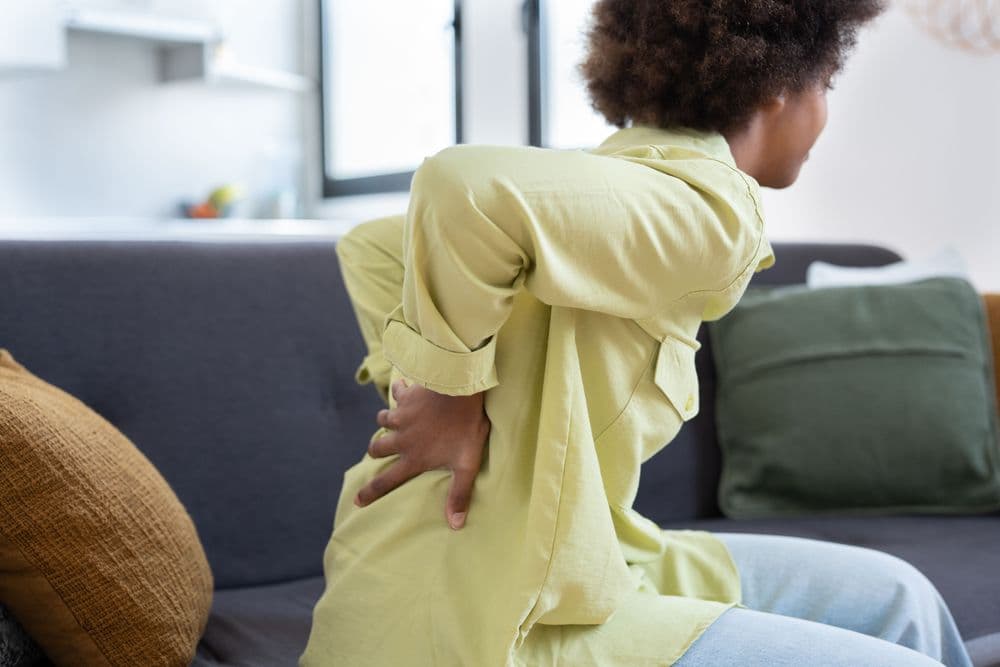 Pessoa jovem, com expressão de desconforto, sentada em um sofá enquanto segura a região lombar com as duas mãos, indicando dor nas costas.