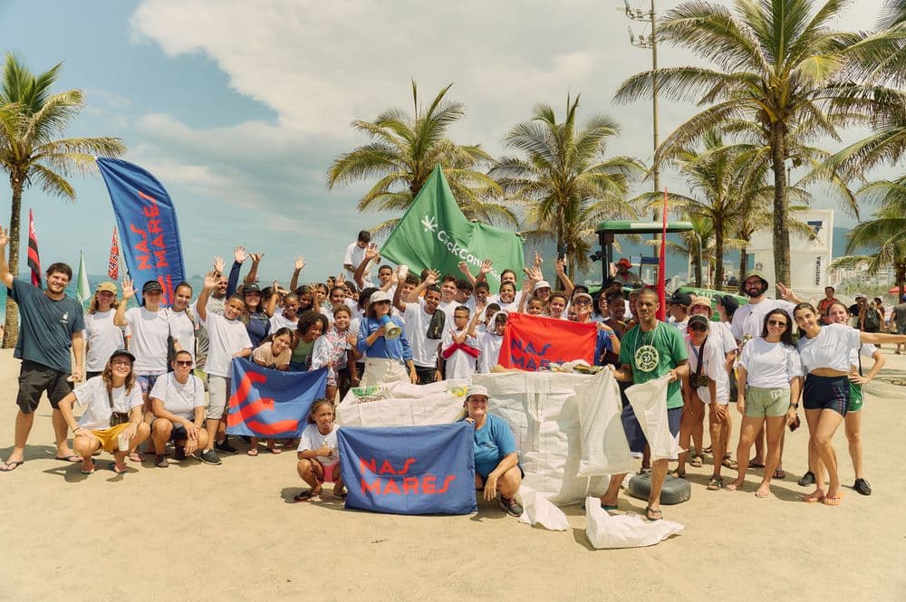 Grande grupo de voluntários participando de um mutirão de limpeza de praia. Eles seguram sacos cheios de lix, além de bandeiras com os nomes das iniciativas envolvidas.