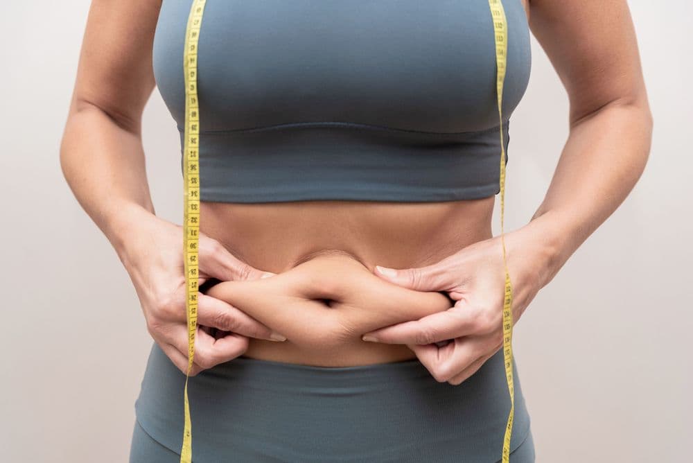 Mulher usando roupa de ginástica azul e segurando a barriga, evidenciando a gordura abdominal, enquanto uma fita métrica amarela passa pelo corpo.