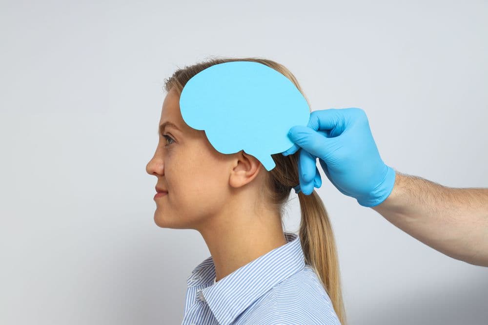 Mulher de perfil, usando camisa listrada azul, enquanto uma mão com luva azul segura um recorte de papel azul em forma de cérebro próximo à sua cabeça.