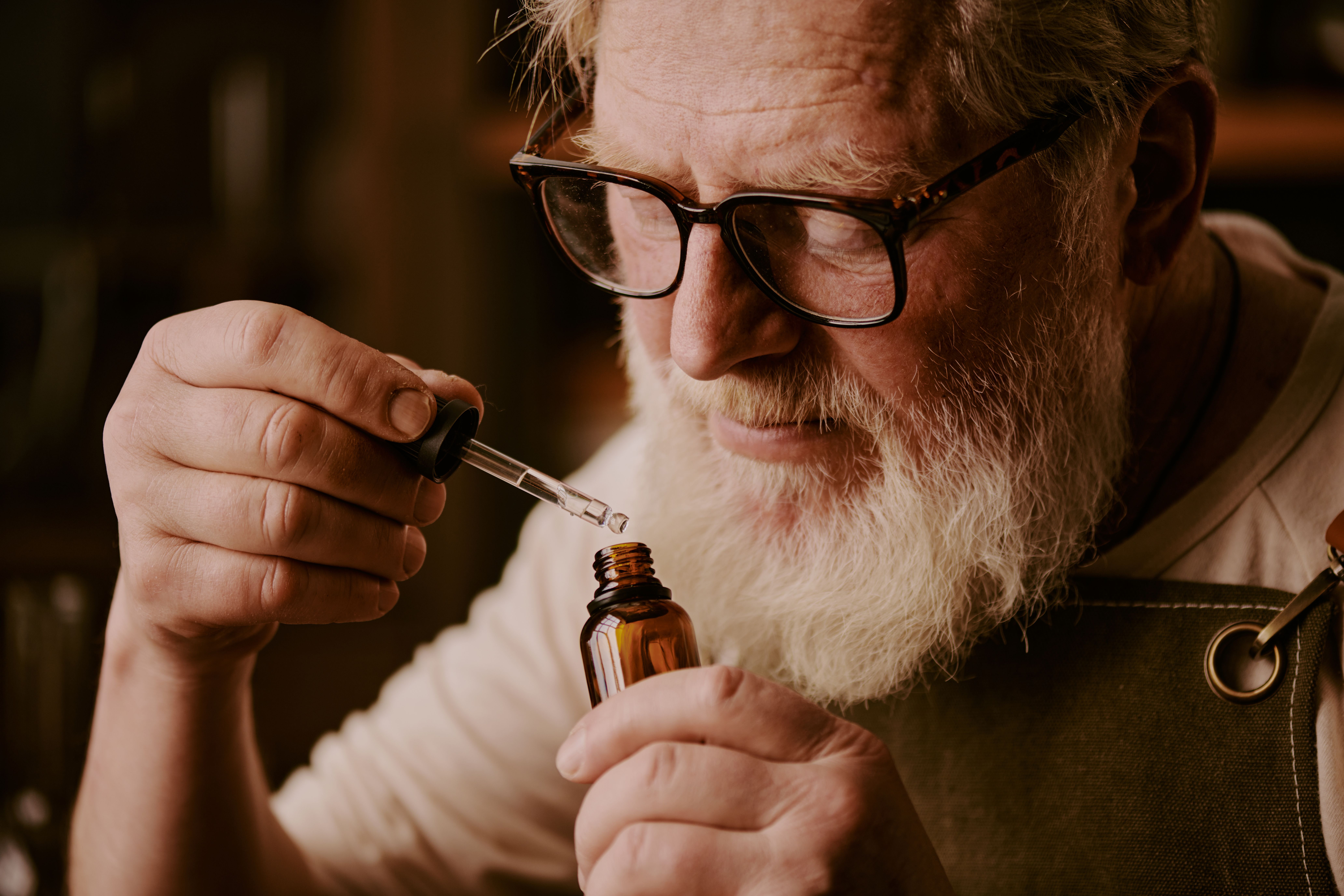 Homem idoso com barba branca e óculos segurando um frasco de vidro âmbar e um conta-gotas com óleo de canabidiol.