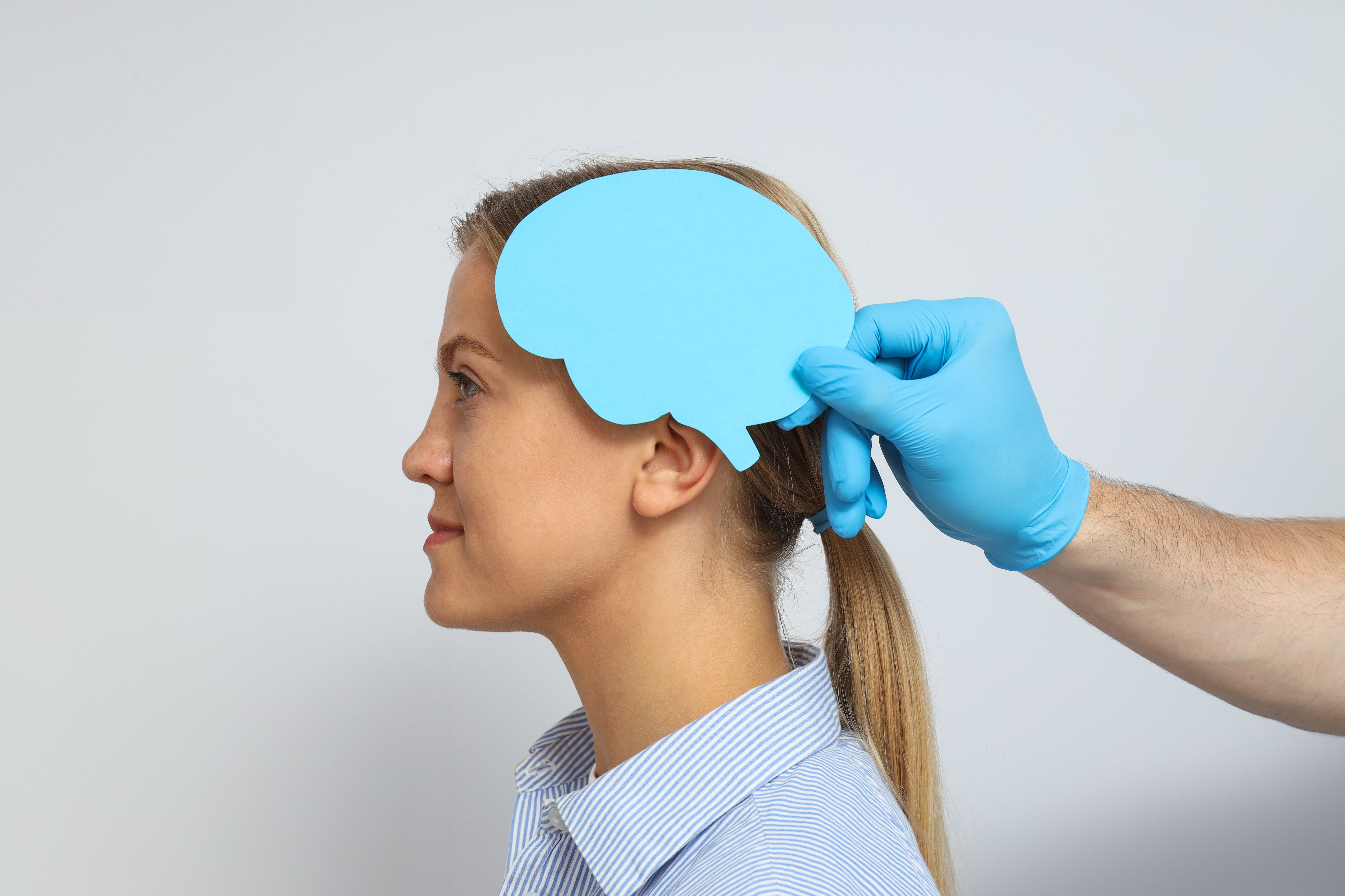 Mulher de perfil, usando camisa listrada azul, enquanto uma mão com luva azul segura um recorte de papel azul em forma de cérebro próximo à sua cabeça.