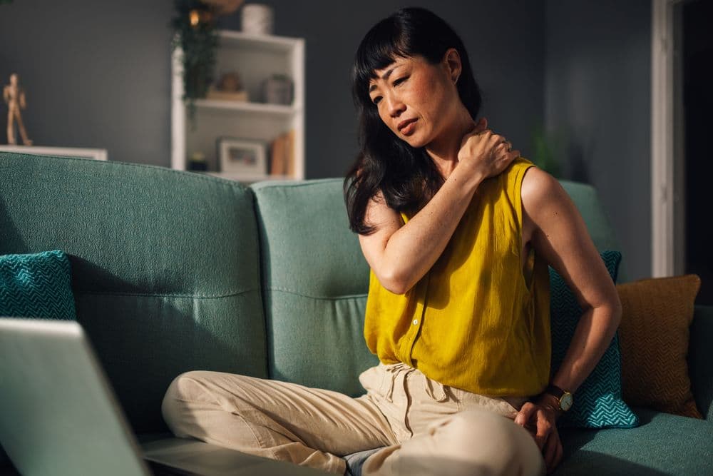Mulher com dor no ombro sentada no sofá, sinal de inflamação crônica
