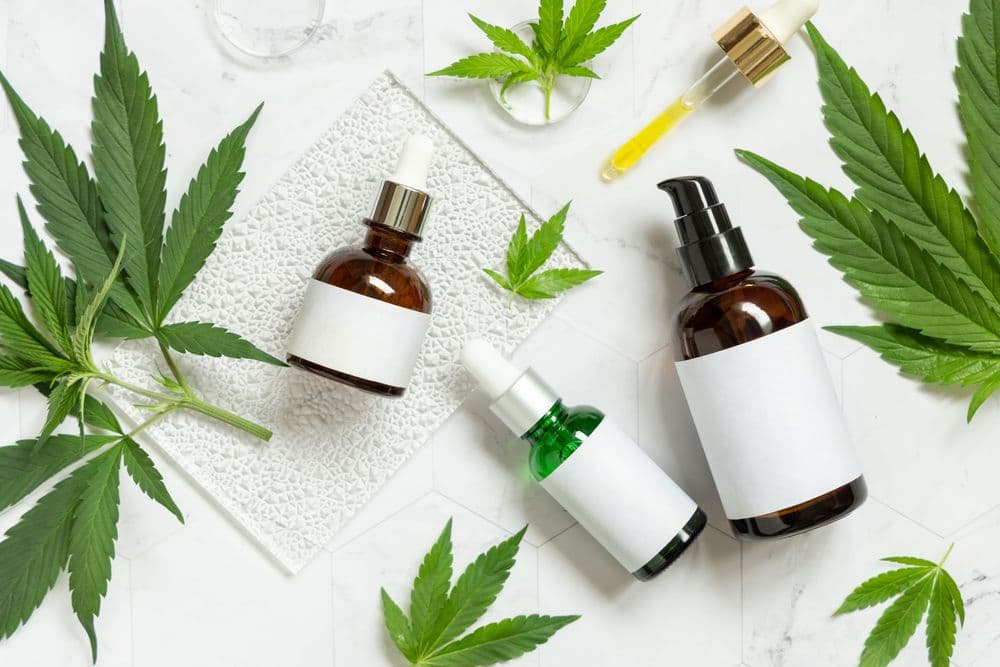 Frascos de óleos e produtos tópicos à base de cannabis ao lado de folhas de cannabis sobre superfície branca.