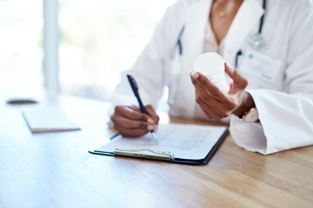 Médica segurando um frasco de medicamento enquanto escreve uma prescrição em consulta clínica