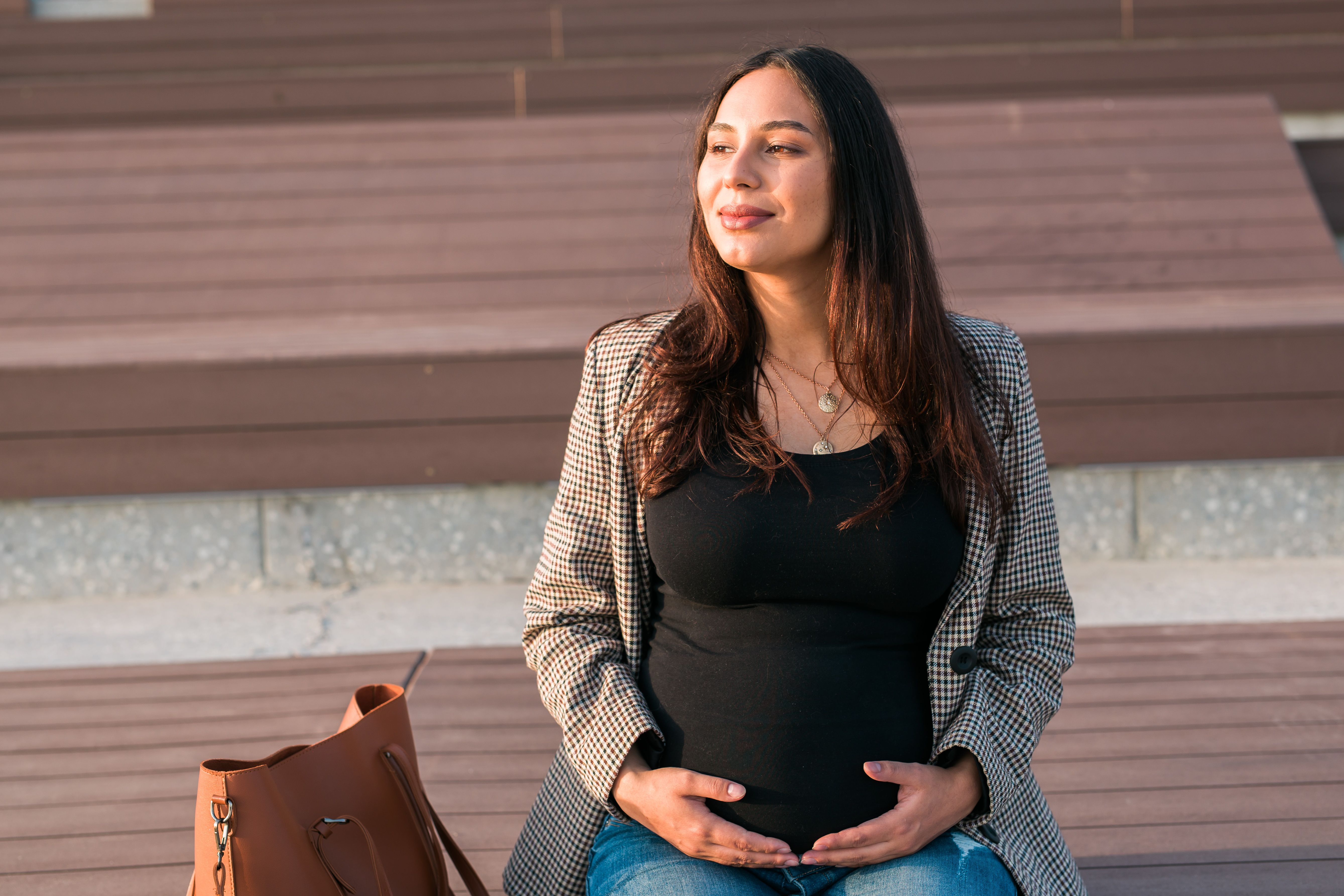 Mulher grávida sentada ao ar livre, sorridente, com as mãos sobre a barriga, aproveitando a luz do sol.