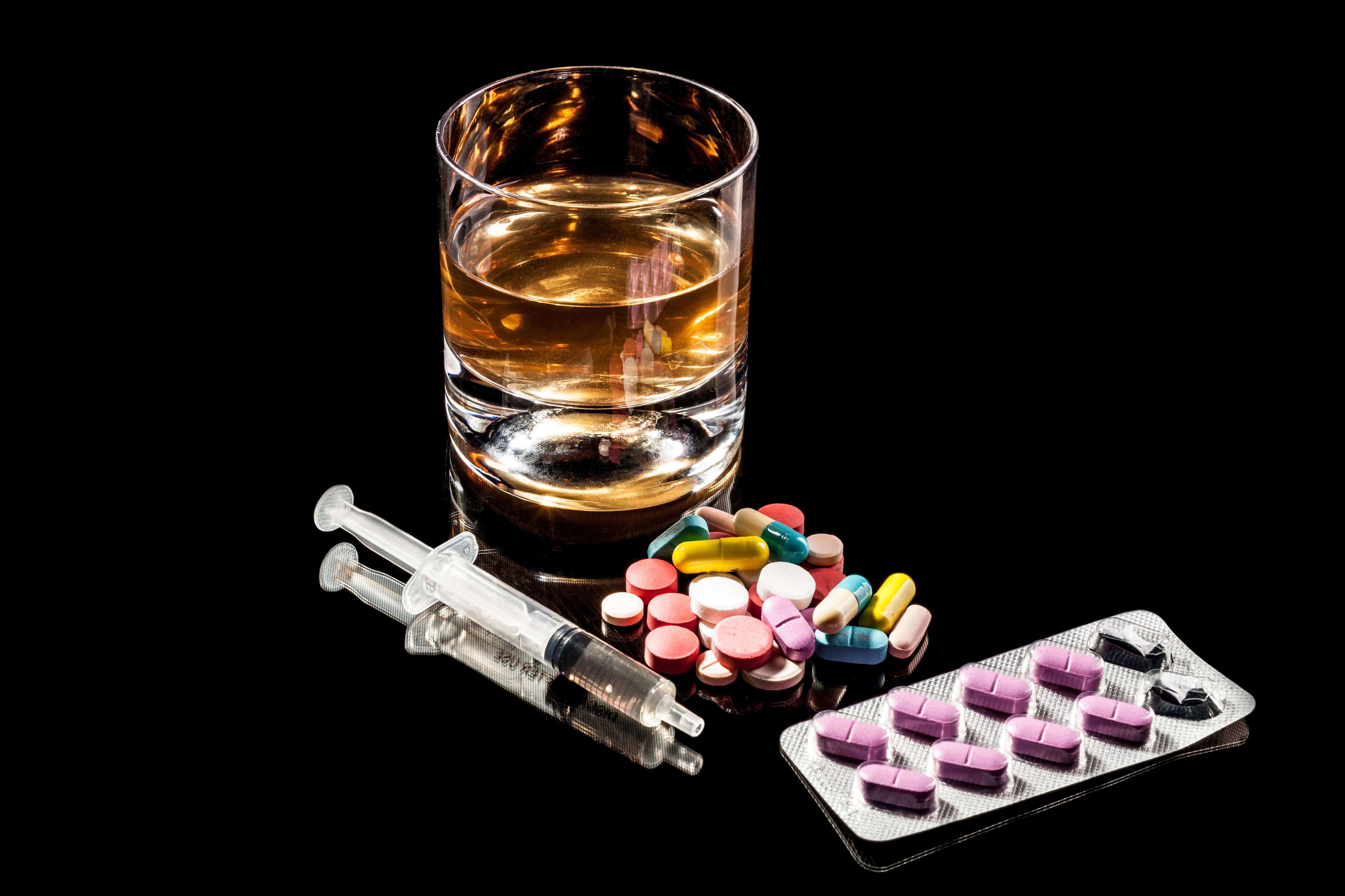  Copo com bebida alcoólica em destaque sobre fundo preto, acompanhado por duas seringas, diversos comprimidos coloridos soltos e uma cartela de medicamentos.