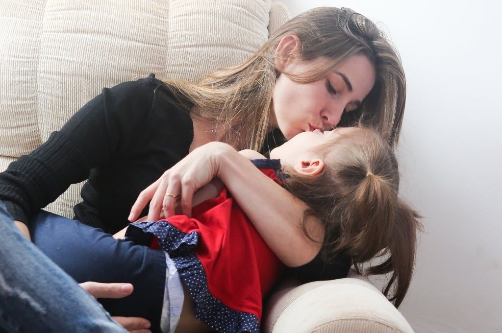 Mãe beijando a filha no colo sentada em um sofá. 