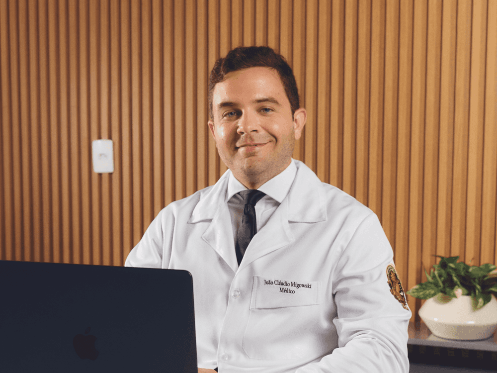Médico sorrindo em consultório, vestindo jaleco branco e gravata, sentado diante de um notebook com fundo de madeira