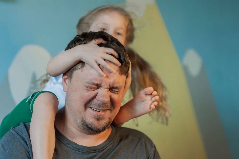 Homem com expressão de incômodo enquanto filha pequena o abraça por trás, representando um momento de descontrole típico de tiques involuntários.