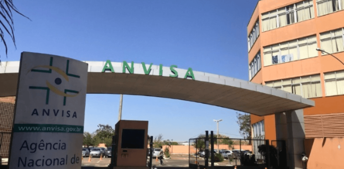 Entrada da sede da Anvisa em Brasília com a placa institucional e o arco com o nome da Agência sob céu azul.