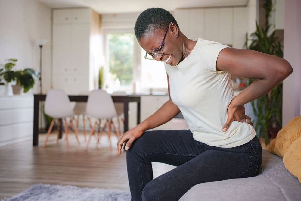 Mulher sentada no sofá em casa segurando a região lombar, demonstrando dor nas costas.