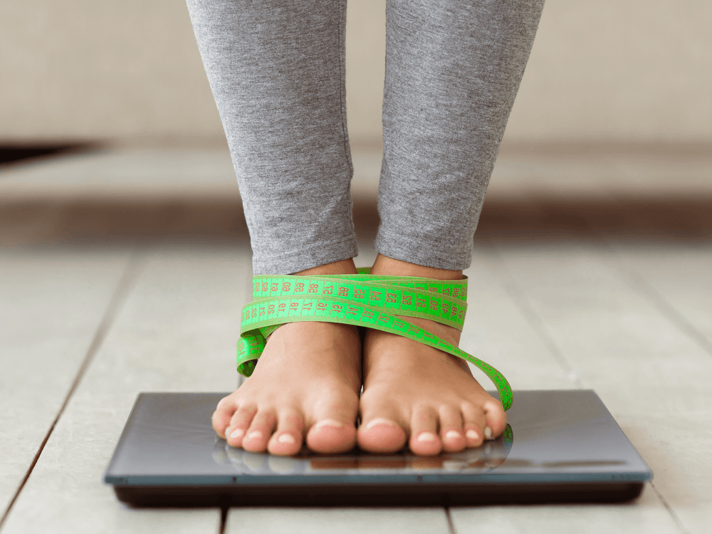 Pessoa em balança digital com fita métrica verde enrolada nos pés, representando controle de peso e metabolismo.