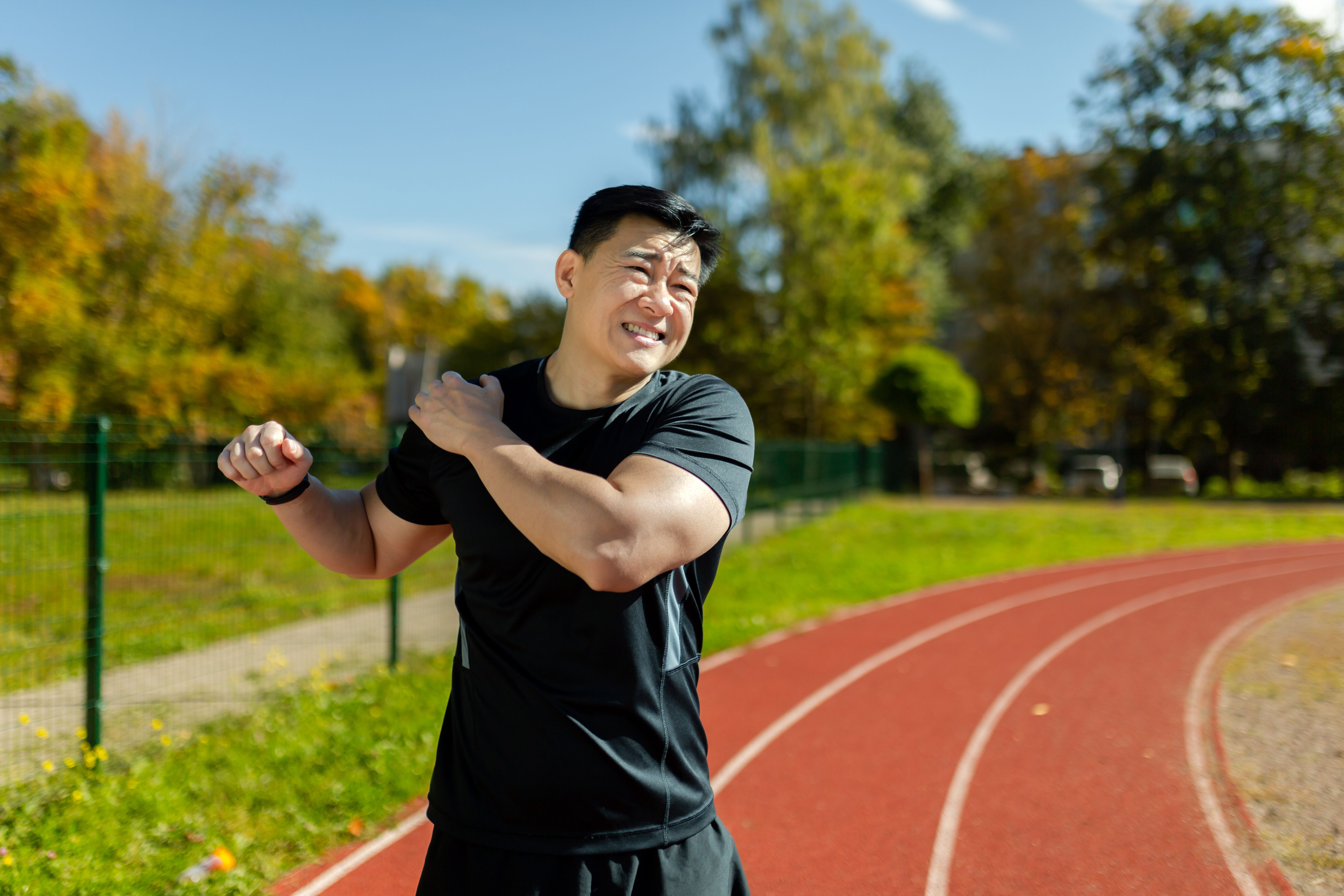 Homem asiático vestindo roupas esportivas pretas, em uma pista de corrida ao ar livre durante o dia com desconforto no ombro direito.