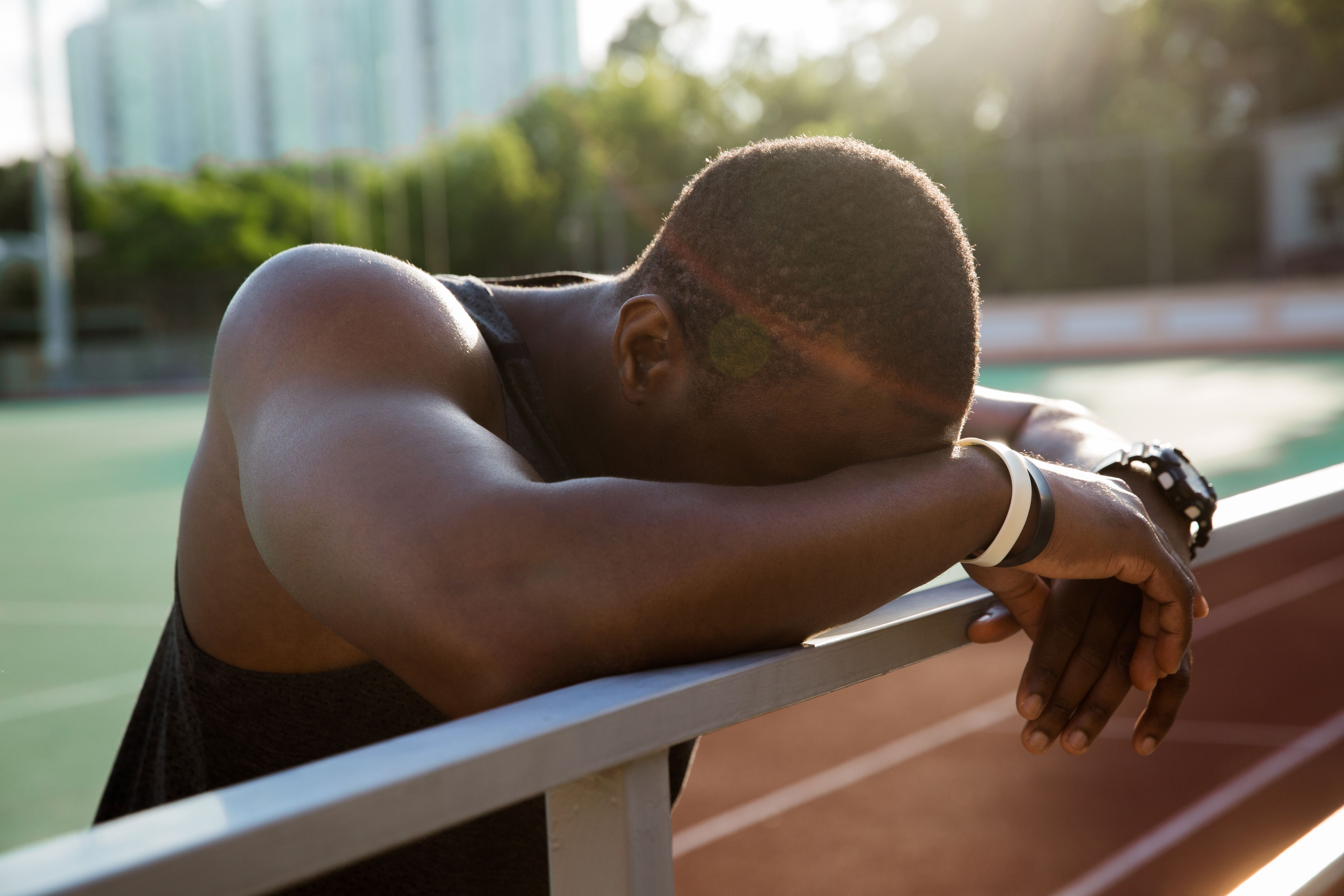 Atleta descansando sobre uma grade ao lado de uma pista esportiva, com o corpo inclinado para frente e a cabeça apoiada nos braços, demonstrando sinais de exaustão física.