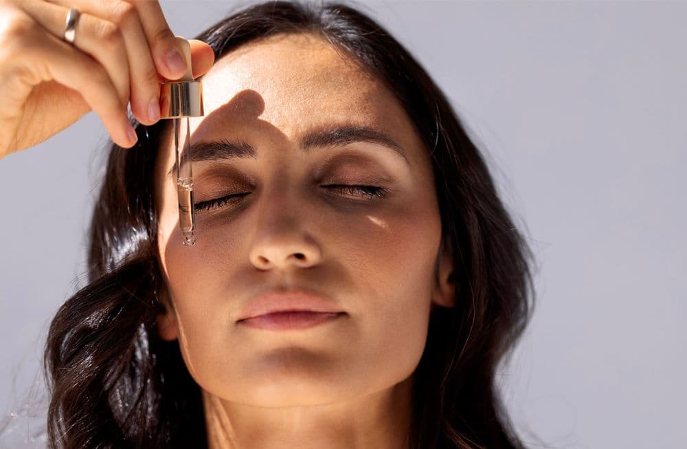 Mulher com olhos fechados aplicando óleo de CBD no rosto para cuidados com a pele