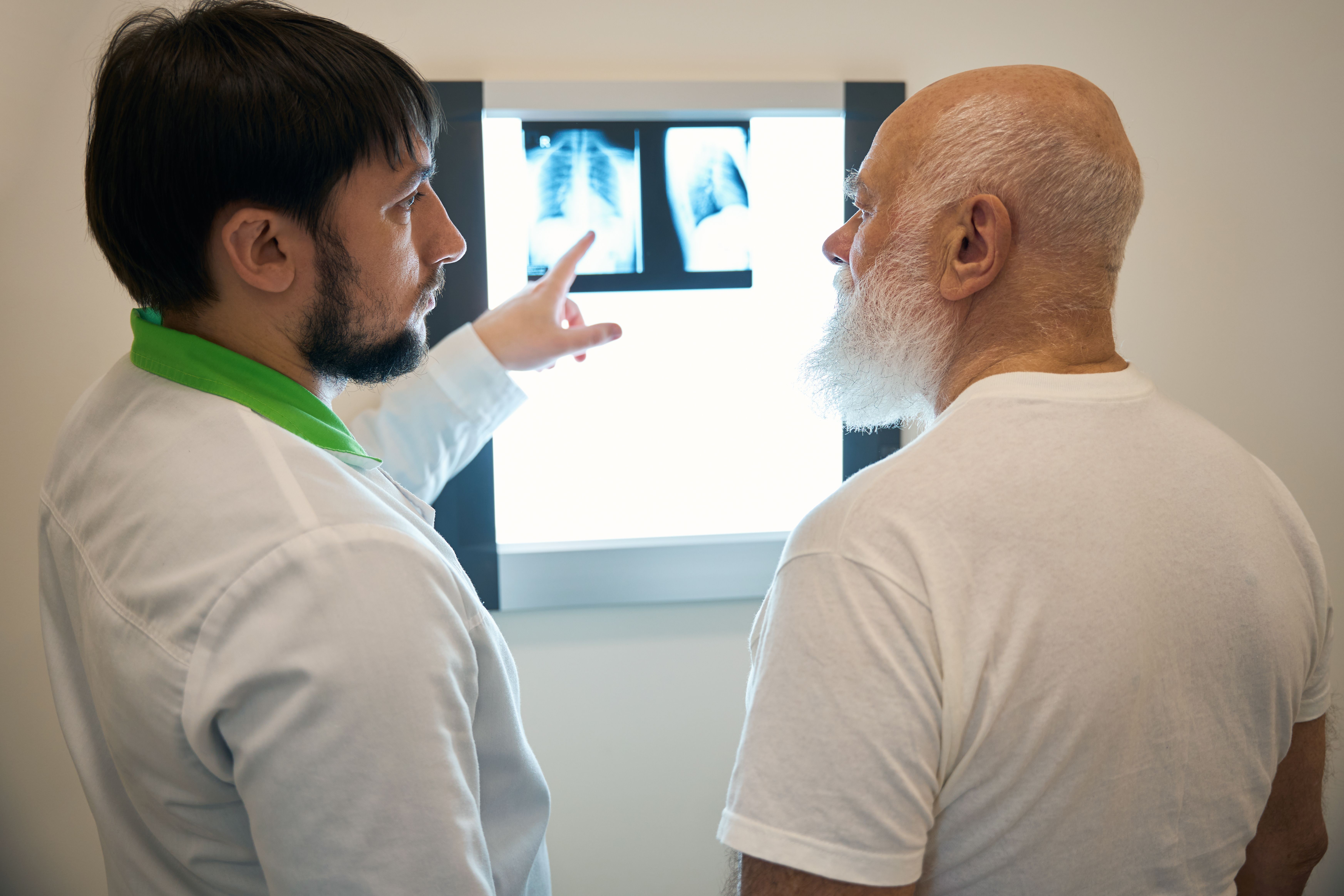 Médico com barba curta e cabelos escuros, usando jaleco branco, apontando para radiografias em um painel de luz enquanto conversa com um paciente idoso, calvo e com barba branca.