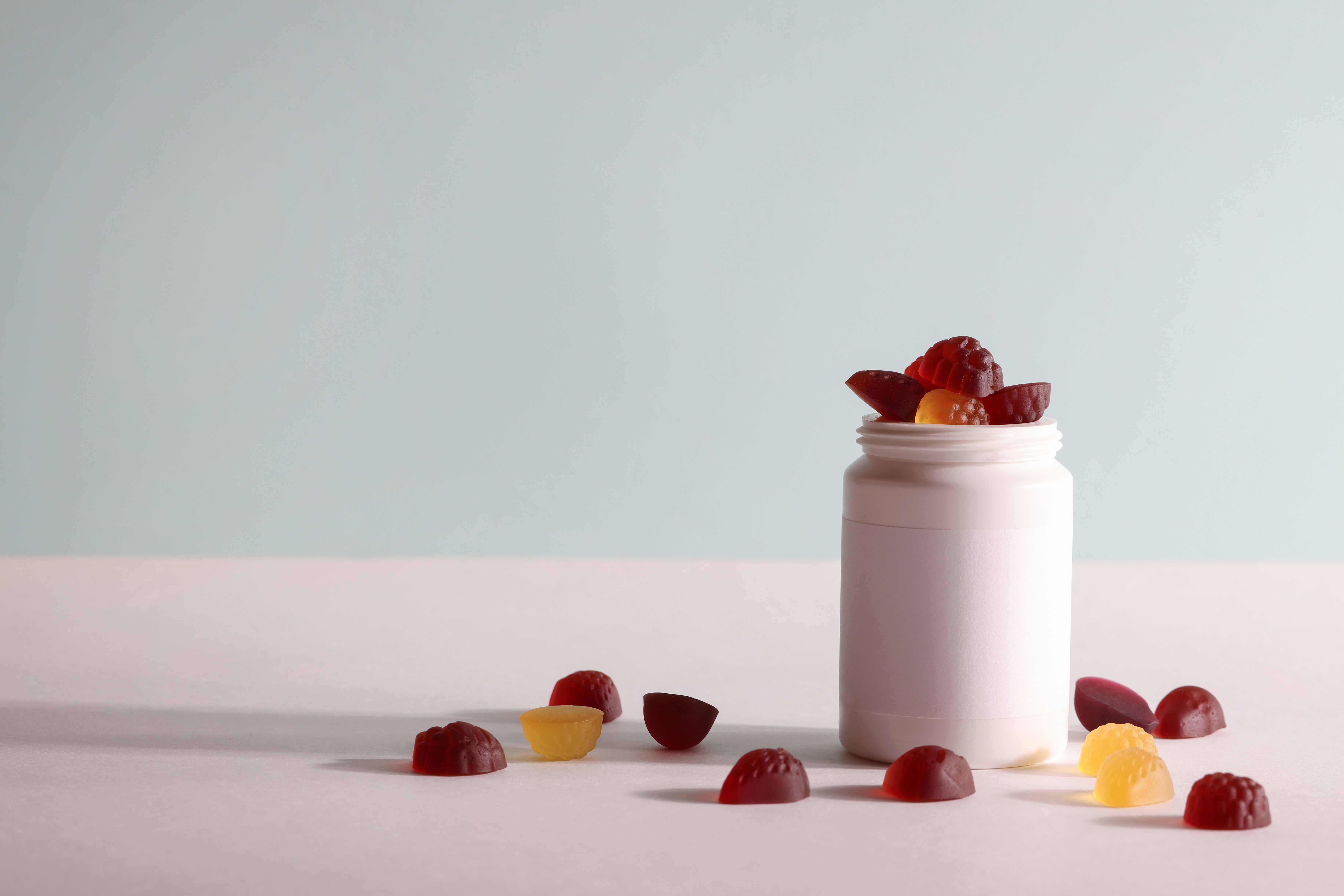 Frasco branco aberto contendo balas de goma coloridas, com algumas espalhadas sobre superfície clara.